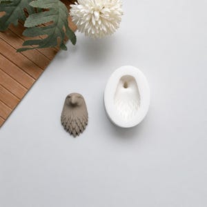Può includere: Un oggetto artigianale a forma di gufo grigio con un design di piume strutturate, accanto a uno stampo in silicone bianco della stessa forma. Il gufo è su una superficie grigio chiaro, con un fiore bianco e foglie verdi sullo sfondo. Artigianato e fai da te.