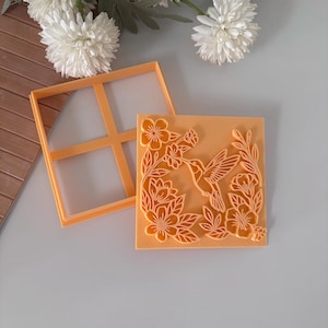 May include: An orange square cookie cutter with a hummingbird and floral design. The cutter is next to a matching frame. The design is detailed with a hummingbird in flight surrounded by flowers and leaves. The background is a light orange color.