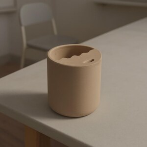 Puede incluir: Un vaso de cer&aacute;mica beige con un dise&ntilde;o ondulado &uacute;nico en el borde. La taza cil&iacute;ndrica tiene un acabado liso y mate y un peque&ntilde;o agujero cerca del borde. El vaso est&aacute; sobre una superficie de color claro.