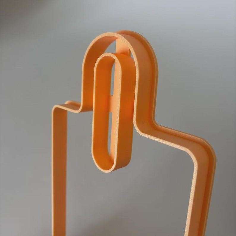 May include: A light orange, abstract, 3D-printed object with a curved, geometric design. The object has a looped top section and a long, straight vertical section. The design is modern and minimalist, suitable for home decor or artistic display.