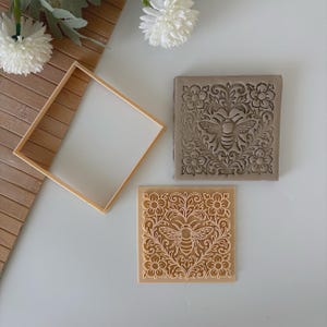 May include: Two square decorative molds with a bee and floral design. One is light brown, the other is gray. A square wooden frame is also in the image. The molds are likely for crafts or home decor.