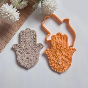 May include: A beige, intricately carved Hamsa hand cookie and an orange cookie cutter. The Hamsa hand features an eye, crescent moons, and floral designs. The cookie cutter is in the shape of the Hamsa hand. The items are on a white surface.