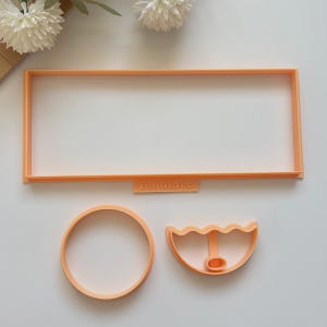 May include: Three orange plastic cookie cutters in various shapes. One is a rectangle, another is a circle, and the third is a half-circle with a wavy edge. The word "mouldis" is printed on the rectangle cutter.