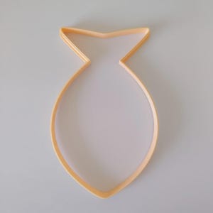 May include: A light orange plastic cookie cutter in the shape of a fish. The cutter has a rounded body and a pointed tail and head. The cutter is on a white background.