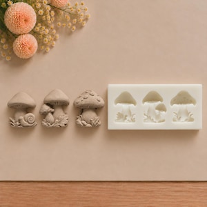 May include: A white rectangular silicone mold featuring three distinct mushroom-themed cavities, accompanied by three finished light brown mushroom-shaped figures, each displaying unique details like a snail, leaves, and spots.