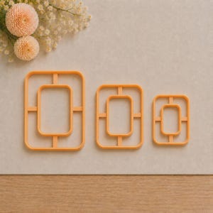 Può includere: Tre tagliabiscotti rettangolari arancioni di diverse dimensioni, ciascuno caratterizzato da un design a doppia parete con quattro piccole barre di collegamento tra il rettangolo interno ed esterno, disposti orizzontalmente.