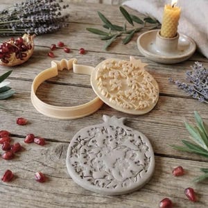 Può includere: Un set di tagliapasta e timbro a forma di melograno. Il timbro presenta un design floreale dettagliato. Il set è beige e grigio, con semi di melograno sparsi e rametti di lavanda su una superficie di legno.