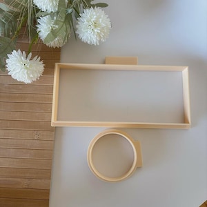 Può includere: Una cornice rettangolare e un telaio da ricamo rotondo beige chiaro, entrambi in legno, sono esposti su una superficie bianca. Fiori bianchi artificiali e un tappetino di bambù sullo sfondo. La cornice ha un piccolo pezzo rettangolare attaccato in alto.