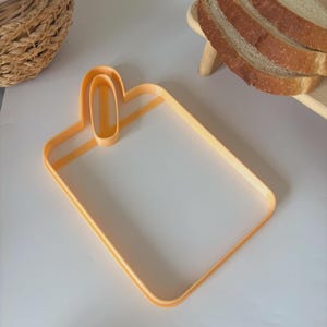 Può includere: Un tagliabiscotti arancione a forma di pane con un manico. Il tagliabiscotti è rettangolare con angoli arrotondati e un manico nella parte superiore. Pane a fette visibile sullo sfondo.