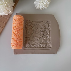 May include: An orange clay roller with a detailed dragon design next to a clay slab with a matching dragon relief. The dragon is surrounded by clouds and decorative elements. The roller is cylindrical.
