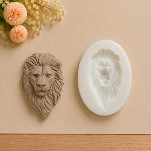 Peut inclure: Une moulage de tête de lion en trois dimensions de couleur brun clair à côté d'un moule en silicone blanc de forme ovale présentant un motif de tête de lion en creux identique. La tête de lion montre des traits détaillés et une crinière texturée.