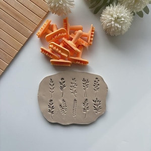 May include: A collection of orange clay stamps with floral designs, alongside a piece of clay imprinted with the same patterns. The stamps have a handle and a textured base. The clay is a light brown color.