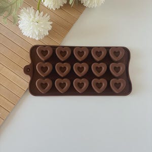 Op de afbeelding: Een bruine siliconen mal met 18 hartvormige holtes. De mal heeft een rechthoekige vorm met een klein handvat aan de linkerkant. De harten zijn gelijkmatig verdeeld, ideaal voor het maken van chocolade.
