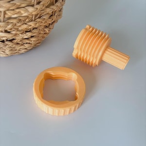 Puede incluir: Un juego de cortadores de galletas naranja impresos en 3D con una base redonda y estriada y un centro en forma de oso. También hay un rodillo a juego con un barril estriado. Una cesta tejida está en el fondo.