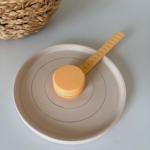 May include: A light orange measuring tool rests on a light pink plate with concentric black circles. The measuring tool has a circular base and a long, narrow handle. A woven basket is partially visible in the background.