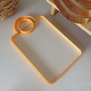 Op de afbeelding: Een oranje plastic broodsnijder met een rechthoekig snijvlak en een cirkelvormige handgreep. De snijder is ontworpen om brood op zijn plaats te houden tijdens het snijden. Gesneden brood is zichtbaar op de achtergrond.