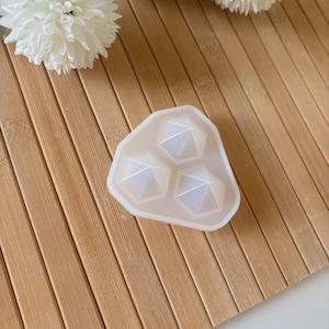 Peut inclure: Un moule en silicone blanc avec trois formes de pyramides géométriques. Le moule est posé sur une surface en bois, avec un design simple et moderne. Le moule est conçu pour le bricolage.