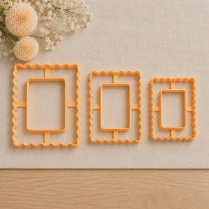 May include: Three light orange rectangular cookie cutters of varying sizes, each featuring a scalloped outer edge and a smooth inner rectangular frame connected by four thin support bars, arranged horizontally from largest to smallest.