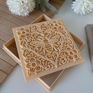May include: A square, beige-colored stamp with a detailed bee and floral design. The stamp is set in a matching box, with the intricate pattern raised from the surface. The design includes a bee surrounded by flowers and leaves.
