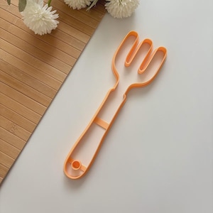 May include: An orange plastic fork-shaped cookie cutter. The cutter has three tines and a long handle with a small circular hole at the end. The cutter is on a white surface with a wooden surface and white flowers in the background.