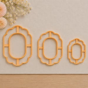 May include: Three orange plastic cookie cutters in graduated sizes, each featuring a scalloped outer frame and a smooth oval inner centre connected by four thin support bars, arranged horizontally from largest to smallest on a flat surface.