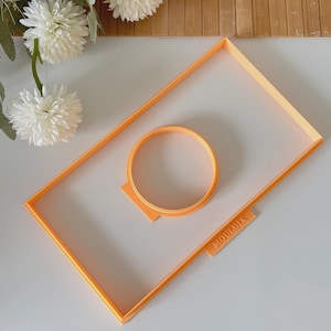 May include: Orange plastic cookie cutters in the shape of a rectangle and a circle. The rectangle cutter has a small tab with the word "MOULDIX" printed on it. The cutters are on a white surface.