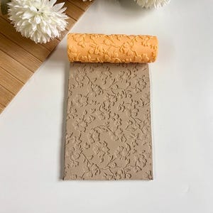 Op de afbeelding: Een oranje deegroller met bloemenpatroon ligt op een beige, getextureerd oppervlak met een bijpassend bloemmotief. De deegroller en het oppervlak zijn ontworpen voor het creëren van reliëfpatronen op materialen zoals klei of deeg.