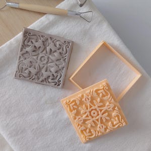 Puede incluir: Dos baldosas de cerámica cuadradas con diseños florales, una gris y otra melocotón, sobre un paño blanco. Un utensilio de alfarería con mango de madera y una herramienta de bucle metálico en el fondo. Las baldosas son probablemente para manualidades o decoración.