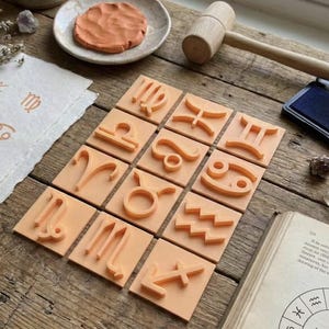 Op de afbeelding: Een verzameling van twaalf vierkante, oranje dierenriemstempels, gerangschikt op een houten oppervlak. Een houten hamer, een inktkussen en een klein schaaltje met klei zijn ook aanwezig. De stempels hebben verhoogde symbolen.