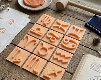 May include: A collection of twelve square, orange-colored zodiac sign stamps arranged on a wooden surface. A wooden mallet, ink pad, and a small dish with clay are also present. The stamps feature raised symbols representing astrological signs.
