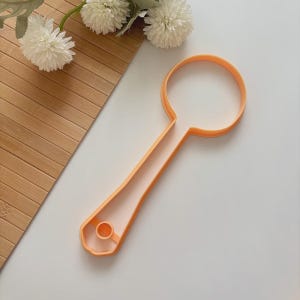 May include: An orange plastic spoon-shaped cookie cutter. The cutter has a circular head and a long, flat handle with a small hole at the end. The cutter is on a white surface with a wooden background and white flowers.