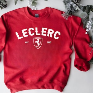 May include: A red sweatshirt with the name "LECLERC" in white block letters. Below the name is a white shield with a prancing horse logo and the text "EST 1997". The sweatshirt has long sleeves.
