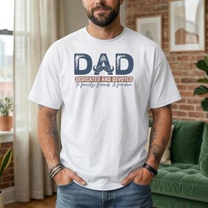 May include: White t-shirt with the word "DAD" in a distressed blue font, a mustache graphic, and the text "Dedicated and Devoted To family, friends & freedom" in a red font. The shirt is a casual style.