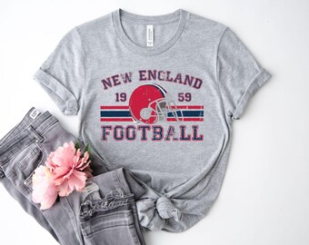 May include: A heather gray t-shirt with a distressed graphic that reads "NEW ENGLAND FOOTBALL 1959." The design includes a red football helmet and red and blue stripes. The shirt is paired with gray jeans and a pink flower.
