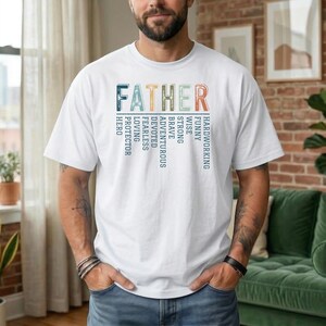 May include: White t-shirt with the word "FATHER" in colorful block letters. Vertical text on the left and right sides lists qualities like "HERO" and "HARDWORKING". The shirt is worn by a person with a beard and tattoos.