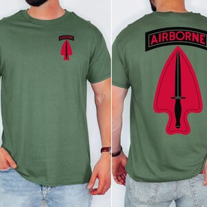 May include: Olive green t-shirt with a red and black design. The front features a red arrow with a black knife and the word "AIRBORNE" in a black rectangle. The back of the shirt has the same design, but larger.