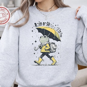 May include: A light gray crewneck sweatshirt featuring a graphic of a frog wearing a yellow jacket and boots, carrying a backpack and a yellow umbrella. The text "Rain or Shine I'll be fine" is printed above the frog, surrounded by small star icons.