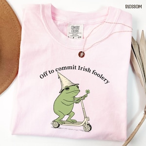 May include: A light pink t-shirt with the text "Off to commit Irish foolery" above a cartoon frog riding a scooter and holding a four-leaf clover. The shirt has a Comfort Colors tag and a gold necklace.