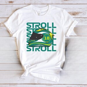 May include: White t-shirt featuring a green and yellow racing helmet graphic with the number 18. The word "STROLL" is printed vertically on the left side and below the helmet. The shirt is made of a soft material.
