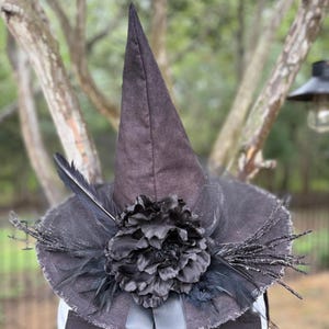 May include: A pointed black witch hat featuring a large black fabric flower at the center, accented with black feathers and glittery black twigs. The hat has a wide, frayed brim and a smooth black ribbon hanging down from the base of the crown.