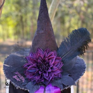 May include: A black pointed hat featuring a large, deep purple dahlia flower at the center, a long black feather, and a frayed brim. The text "Witching Hour" is embroidered in pink on the brim, and a purple ribbon is tied at the base.