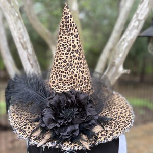 May include: A pointed witch hat featuring a tan and black leopard print pattern. The hat is decorated with a large black fabric flower, black feathers, and black tulle netting around the base. A black apron with the text MOONSPELL HOLLOW is visible.