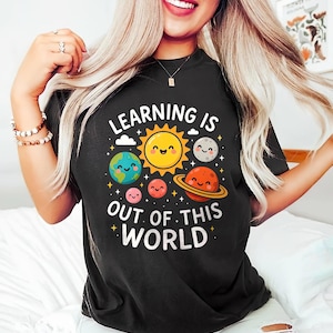 以下が含まれることがあります： 黒いTシャツ。カラフルな惑星のグラフィック、笑顔の太陽、白い文字で「LEARNING IS OUT OF THIS WORLD」と書かれています。デザインは漫画の惑星と星を特徴とし、楽しく教育的なテーマを作成しています。