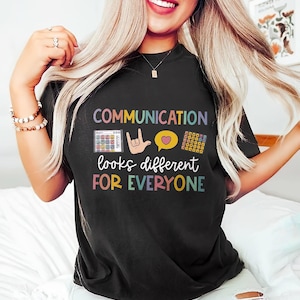以下が含まれることがあります： カラフルな文字で「COMMUNICATION looks different FOR EVERYONE」と書かれた黒いTシャツ。コミュニケーションボード、手のサイン、ハートの吹き出しの画像が含まれています。
