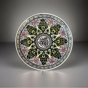 May include: A round ceramic plate with an intricate floral design. The plate features a central medallion with Arabic calligraphy, surrounded by green, black, yellow, red, and blue floral patterns. The plate has a white border.