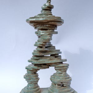 May include: A teal ceramic sculpture with a layered, abstract design. The sculpture features a base with two stacked columns supporting a central archway. The top has a spiraling, tiered structure. The piece is approximately 10 inches tall.