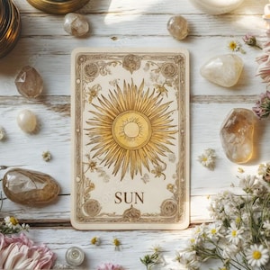 Puede incluir: Una carta de tarot con un diseño de sol, rodeada de cristales, flores y una vela encendida. La carta presenta una ilustración dorada de rayos de sol con la palabra "SUN" en la parte inferior. La escena está sobre una superficie de madera blanca.