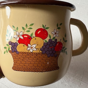 Op de afbeelding: Vintage emaille mok met een beige buitenkant en een bruine rand en handvat. De mok heeft een kleurrijke illustratie van een mand gevuld met rode appels, paarse druiven en gele sinaasappels. Witte bloemen en groene bladeren maken het ontwerp compleet.