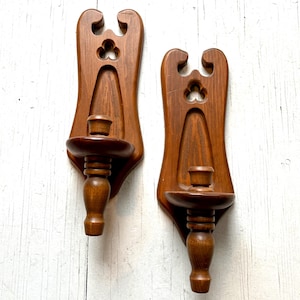 May include: Two wooden wall sconces with a dark brown finish. Each sconce features a decorative cutout at the top and a candle holder. The sconces are mounted on a light-colored, textured surface.