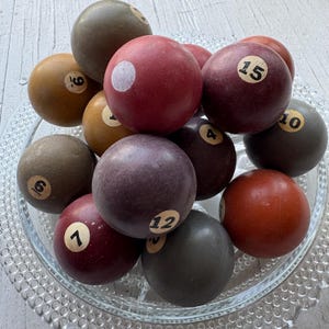 Op de afbeelding: Een verzameling vintage biljartballen in verschillende kleuren, waaronder rood, bruin en groen, met nummers van 1 tot 16. De ballen zijn gerangschikt in een heldere glazen schaal.